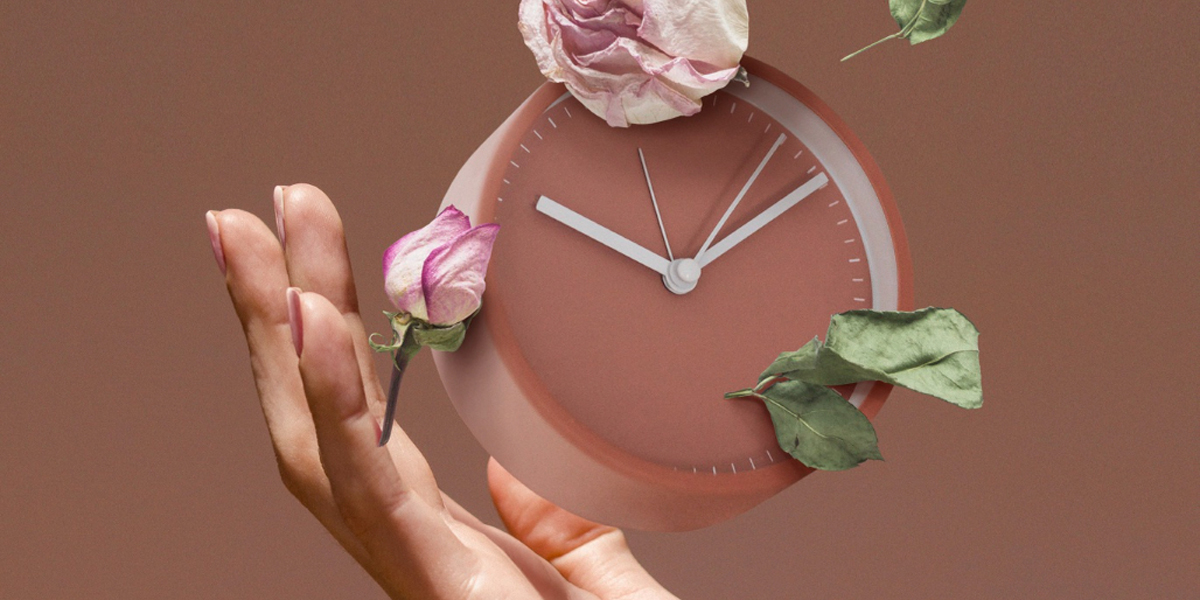 Mano che sostiene un orologio rosa con petali di rosa sospesi intorno, su sfondo beige