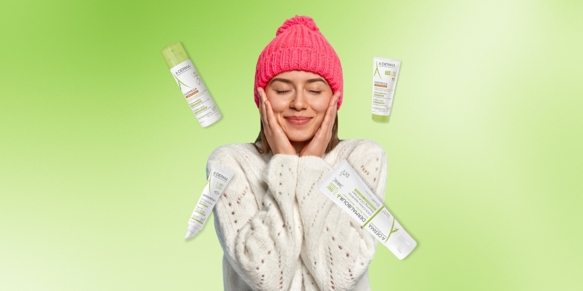 Una donna sorridente con un cappello rosa e un maglione bianco si accarezza il viso, circondata da prodotti della linea A-Derma su uno sfondo verde chiaro. L'immagine comunica una sensazione di benessere e protezione per la pelle durante l'inverno